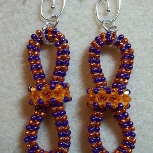 May include: A pair of purple and copper beaded earrings with a silver hook. The beads are arranged in a twisted pattern with a small orange bow in the center of each earring.