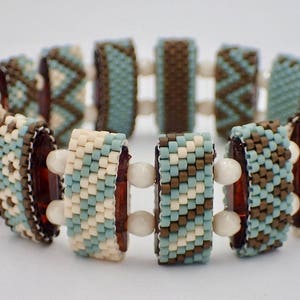 Op de afbeelding: Een kralenarmband met een herhalend patroon van rechthoekige segmenten. De segmenten zijn versierd met kleine, kleurrijke kralen in crème, turquoise en bruin. De armband is verbonden door witte, bolvormige kralen.