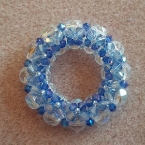 Puede incluir: Una corona de cuentas de cristal azul y transparente, hecha con pequeñas cuentas facetadas. La corona es circular y tiene un aspecto delicado y femenino.