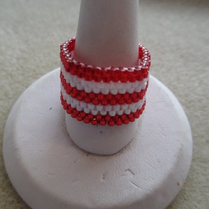 May include: Red and white beaded ring with a striped pattern. The ring is displayed on a white ring stand.