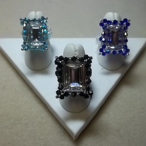 May include: Three rings with clear stones set in a square shape. The rings are surrounded by beads in blue, black, and light blue.