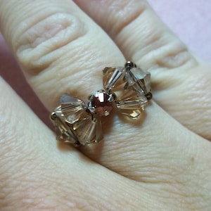 Könnte beinhalten: Ein goldener Ring mit einem braunen Kristall-Schleifendesign. Die Schleife besteht aus mehreren kleinen Kristallen und hat einen größeren, runden, braunen Kristall in der Mitte.