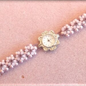 May include: A delicate, light pink beaded bracelet watch with a silver-toned, floral-shaped watch face. The watch band is made of small, round, light pink beads. The watch is set against a soft pink background.