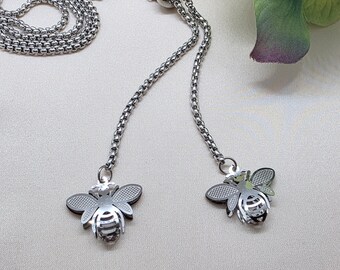 May include: A silver-toned necklace with a delicate chain and two bee-shaped pendants. The bee pendants have detailed wings and body patterns. The necklace is displayed on a neutral background, highlighting the jewelry's design.