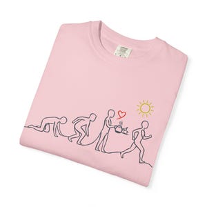 Puede incluir: Camiseta rosa claro con un dibujo lineal negro que representa la evolución de una persona, desde gatear hasta correr. También hay un corazón y un sol dibujados. La camiseta tiene una etiqueta en la esquina superior derecha.