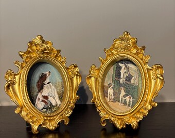 May include: Two ornate, gold-colored oval picture frames. Each frame holds a small, oval-shaped image of dogs. The frames have detailed scrollwork and are displayed on a dark surface.