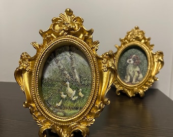 May include: Two ornate, gold-colored oval picture frames. One frame features a painting of ducks, the other a dog. The frames have detailed scrollwork and are displayed on a dark brown surface.