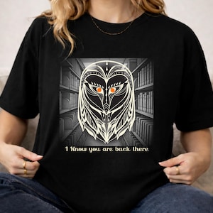 May include: A black t-shirt featuring a graphic print of a stylized owl face with bright orange eyes. The owl is centered against a grayscale background of library bookshelves. Below the owl, the text reads, I Know you are back there.