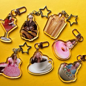 May include: A selection of acrylic keyrings featuring images of a cocktail, a woman in a fur coat, churros, a donut, a woman in a pink dress, a woman in a coffee cup, and cereal. Each keyring has a gold-coloured clasp.