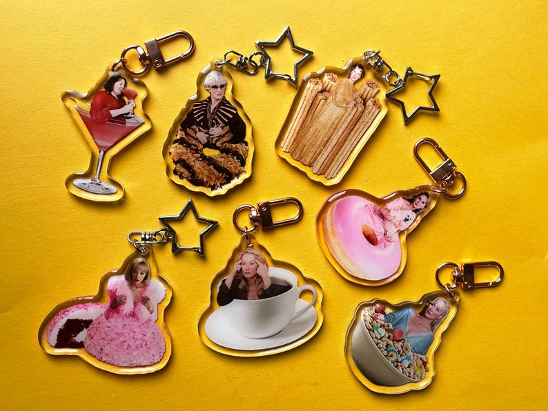May include: A selection of acrylic keyrings featuring images of a cocktail, a woman in a fur coat, churros, a donut, a woman in a pink dress, a woman in a coffee cup, and cereal. Each keyring has a gold-coloured clasp.