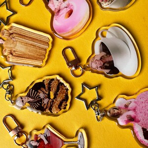 May include: A selection of clear acrylic keyrings featuring images of food and people, such as a donut and a cocktail. Each keyring has a gold-coloured clasp and a silver star-shaped charm. The background is a solid yellow.