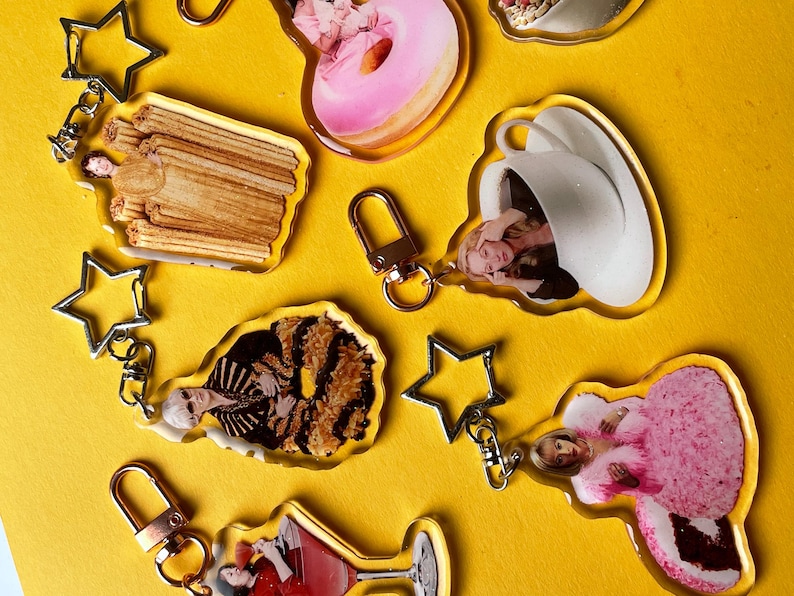 May include: A selection of clear acrylic keyrings featuring images of food and people, such as a donut and a cocktail. Each keyring has a gold-coloured clasp and a silver star-shaped charm. The background is a solid yellow.