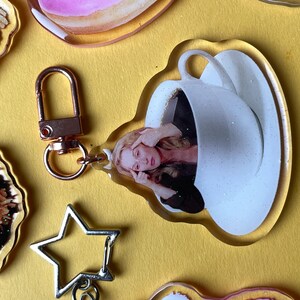 May include: A clear acrylic keyring with a rose gold clasp. The keyring features a woman inside a white teacup. Other acrylic charms in the image include a star, and other decorative shapes.