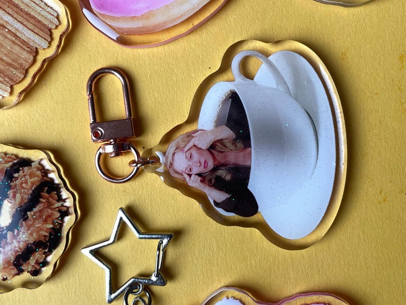 May include: A clear acrylic keyring with a rose gold clasp. The keyring features a woman inside a white teacup. Other acrylic charms in the image include a star, and other decorative shapes.