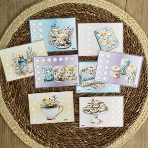 Pode incluir: Uma coleção de cartões em estilo aquarela com ilustrações florais e temáticas de chá. Cada cartão tem um design diferente, incluindo xícaras de chá, velas e arranjos florais, com uma série de pequenos círculos no lado esquerdo. Os cartões estão dispostos sobre um tapete circular tecido.