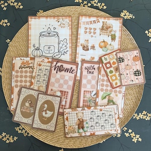 Op de afbeelding: Een verzameling herfstachtige papieren stickervellen en kaarten met illustraties van pompoenen, beren, uilen, ganzen en kaarsen. De ontwerpen bevatten rastertrackers, genummerde cirkels en tekstelementen zoals cozy, home en with love.