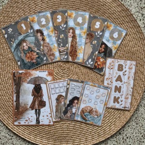 Op de afbeelding: Een verzameling papieren planner-inlays en stickers met herfstthema-illustraties van mensen, een kat en bladeren. De set bevat zes genummerde kaarten, een groter vel met een persoon onder een paraplu en een kaart met het woord BANK in bruine letters.