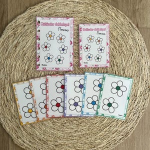 Op de afbeelding: Een set bloemkaarten met de tekst "Multicolor dobbelspel Flowers". De kaarten tonen kleurrijke bloemillustraties met witte bloemblaadjes en verschillende gekleurde centra. De kaarten zijn gerangschikt op een geweven mat.