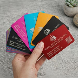 May include: A fan of rectangular metal business cards in red, black, yellow, blue, and pink. Each card features a compass logo, the text TERRA LUMINA SOLUTIONS, Sustainable Consulting, contact details for Alexander Lee, and a QR code.