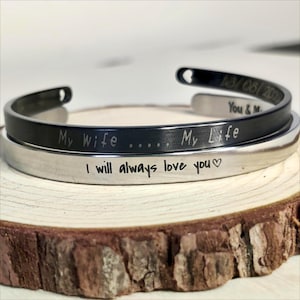Pode incluir: Duas pulseiras de prata com mensagens gravadas. A pulseira superior é preta e está gravada com "My Wife.....My Life". A pulseira inferior é prateada e está gravada com "I will always love you♡".