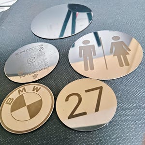 Op de afbeelding: Vijf ronde metalen bordjes met reflecterende oppervlakken. Eén is blanco, één toont een toileticoon, één geeft het getal 27 weer, één toont het BMW-logo en één bevat de tekst SCAN CODE OR TAP TO SEE MENU met bijbehorende iconen.