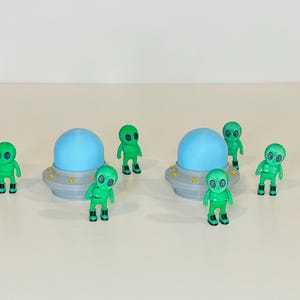 May include: A collection of green alien figurines with black boots and large eyes, alongside two silver and gray UFOs with blue spheres. The toys are arranged on a light-colored surface, creating a playful scene.