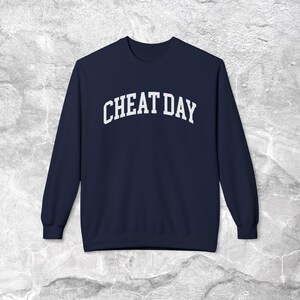 Może przedstawiać: Granatowa bluza z białym napisem "CHEAT DAY" w łukowatym kształcie. Ten top z długim rękawem i okrągłym dekoltem jest wykonany z miękkiego materiału i nadaje się do noszenia na co dzień.