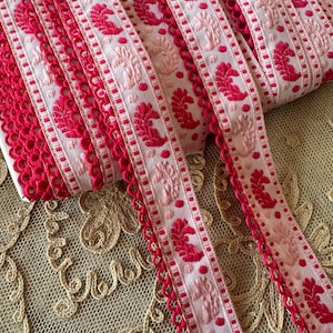 May include: Pink and red woven ribbon trim with a scalloped edge. The ribbon has a repeating pattern of pink and red paisley shapes and dots.