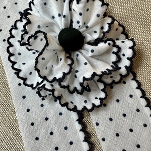 May include: White fabric with black polka dots and a large, ruffled flower. The flower has a black center and scalloped edges. The fabric is cut into strips with scalloped edges.