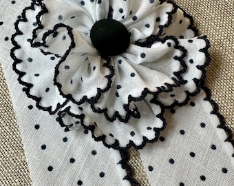 May include: White fabric with black polka dots and a large, ruffled flower. The flower has a black center and scalloped edges. The fabric is cut into strips with scalloped edges.