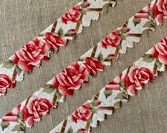 May include: Three strips of white fabric with a red rose pattern and scalloped edges. The fabric is on a brown linen background.