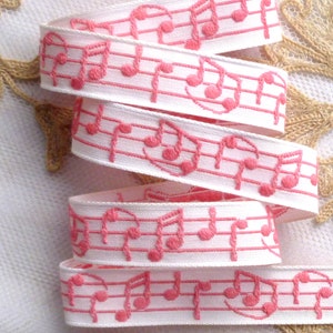 May include: White ribbon featuring embroidered pink musical notes and staff lines. The ribbon is folded, displaying the repeating pattern of musical symbols. Suitable for crafts, sewing, or gift wrapping. The ribbon's width is approximately 2.5 cm.