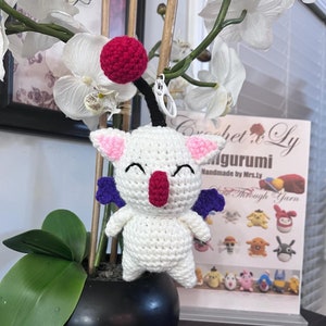May include: White crocheted amigurumi toy with pink ears, purple wings, and a red nose. The toy is sitting on a black pot with green leaves. The background is a white orchid plant and a sign that says "Crochet x Ly Amigurumi Handmade by Mrs.Ly"