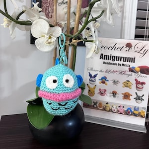 May include: A crocheted blue fish amigurumi keychain with white eyes, a pink mouth, and a blue and white tail. The keychain is hanging from a white orchid plant in a black pot. The background includes a sign that reads "crochet & Ly Amigurumi Handmade by Mrs.Ly Share the Love Through Yarn".