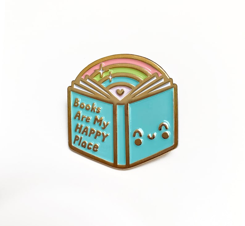 May include: Gold enamel pin shaped like an open book with a rainbow arch above it. The book has a light blue cover with the text "Books Are My Happy Place" and a cute cartoon face.