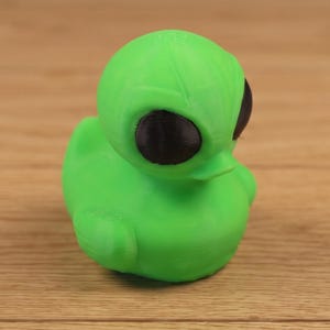 May include: A bright green alien duck figurine with large black eyes. The toy is a whimsical fusion of a classic rubber duck and an extraterrestrial creature. The duck is sitting on a wooden surface.