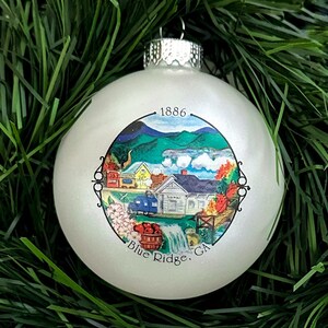 Könnte beinhalten: Weißer Weihnachtsbaumschmuck mit einer bunten Illustration einer kleinen Stadt mit einem Zug, einem Wasserfall und dem Text "1886 Blue Ridge, GA".