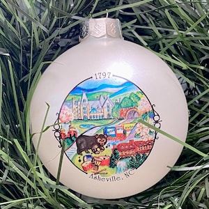 May include: White Christmas ornament with a colorful illustration of Asheville, North Carolina. The ornament features a building, a bridge, a bear, and a waterfall. The text "1797" and "Asheville, NC" are printed on the ornament.