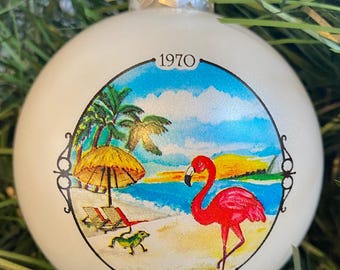 May include: White Christmas ornament with a beach scene featuring palm trees, a beach umbrella, lounge chairs, a pink flamingo, and a green lizard. The ornament has the text "1970 Cancun, Mexico" printed on it.