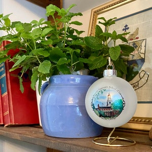 May include: A white glass bauble with a blue and green image of Mercer University School of Law in Macon, Georgia. The bauble is on a gold stand and in front of a potted plant.