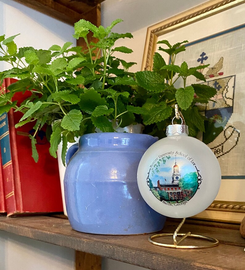 May include: A white glass bauble with a blue and green image of Mercer University School of Law in Macon, Georgia. The bauble is on a gold stand and in front of a potted plant.