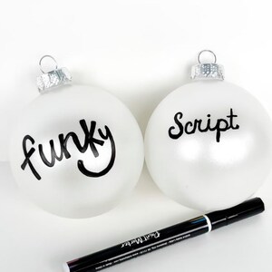 May include: Two white Christmas baubles with black paint marker lettering. One bauble says "funky" and the other says "script". A black paint marker is lying on a white surface.
