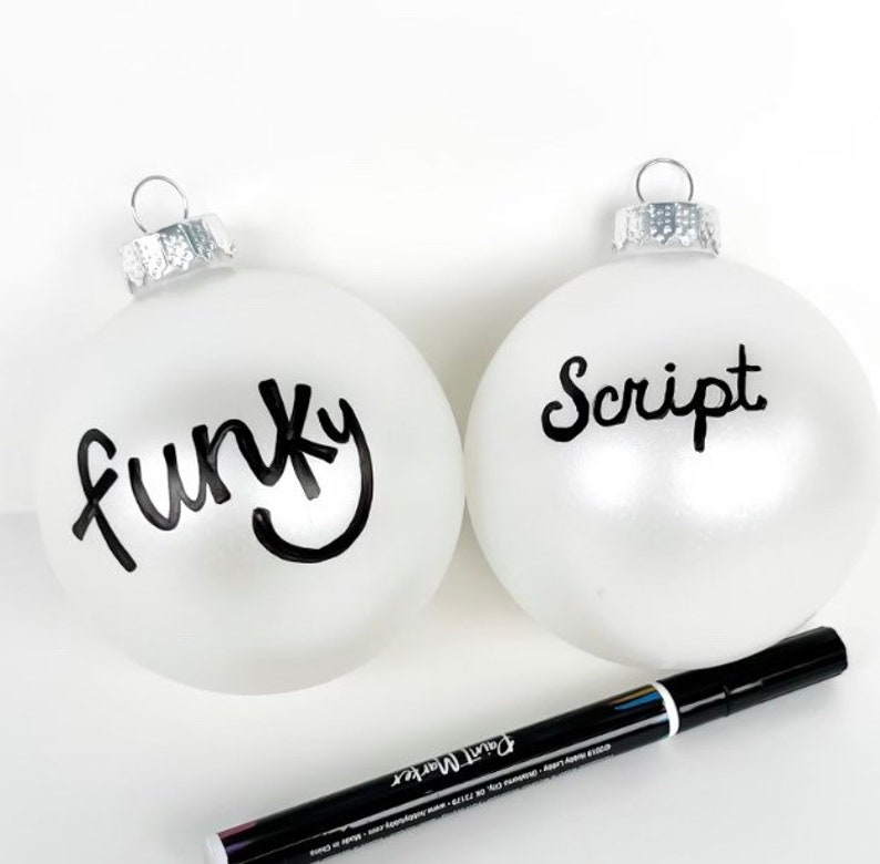 May include: Two white Christmas baubles with black paint marker lettering. One bauble says "funky" and the other says "script". A black paint marker is lying on a white surface.