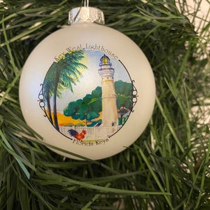 May include: White Christmas ornament with a colorful illustration of the Key West Lighthouse, palm trees, and a rooster. The ornament reads "Key West Lighthouse, Florida Keys".