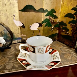 May include: White teacup and saucer set with a geometric pattern in black, red, and gold. The cup and saucer have a gold trim. The background features a gold screen with painted cranes and trees, creating an elegant, Asian-inspired aesthetic.