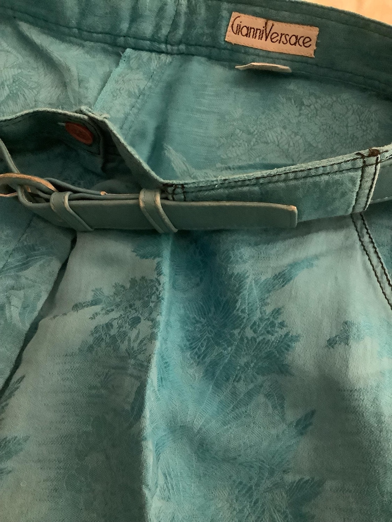 Peut inclure: Un pantalon turquoise bleu avec un motif floral et une ceinture en cuir marron. Le pantalon porte une &eacute;tiquette "Gianni Versace".