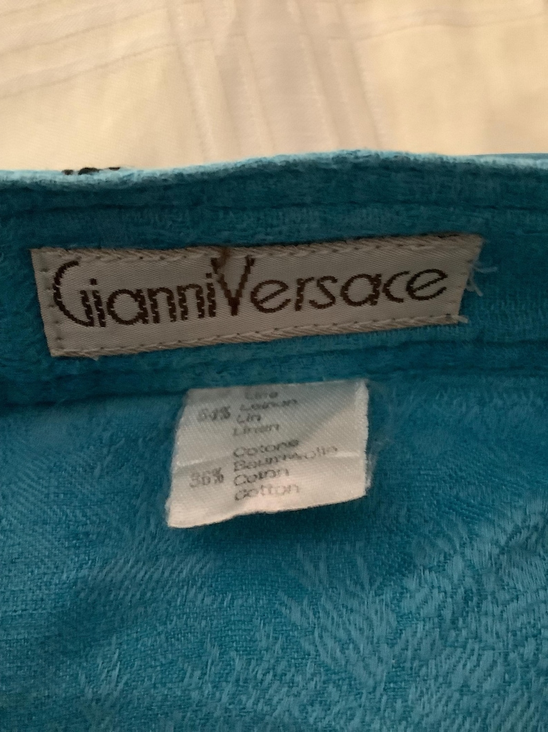 Peut inclure: Un tissu bleu turquoise avec une &eacute;tiquette blanche qui dit "Gianni Versace" et une plus petite &eacute;tiquette blanche avec le texte "Care 64% Linen 36% Cotton Ramie Cotton Cotton".