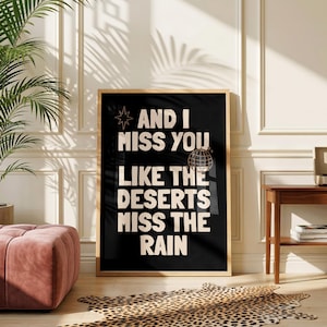 Può includere: Una stampa artistica rettangolare da parete in una cornice di legno chiaro con sfondo nero e testo color crema che recita AND I MISS YOU LIKE THE DESERTS MISS THE RAIN, accompagnata da una piccola icona a stella e una sfera stroboscopica.