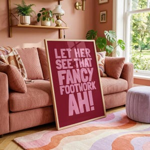 May include: A rectangular framed wall art piece with a dark red background and light pink text that reads: LET HER SEE THAT FANCY FOOTWORK AH! The frame is made of light-coloured wood and the print is positioned in front of a pink corduroy sofa.