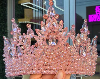 May include: A pink tiara with a sparkling design. The crown is adorned with numerous small, round beads and larger, teardrop-shaped crystals. The tiara is held up by a hand.